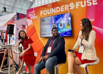 Three people sit on stools and speak on a stage labeled "FOUNDERS FUSE" at London Tech Week. A large screen displays their names and photos behind them inside a spacious, glass-roofed venue. | FintechZoom