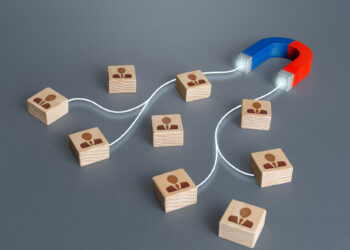 A red and blue horseshoe magnet attracts wooden blocks with person icons on a gray surface, symbolizing customer acquisition or recruiting employees. White lines connect the magnet to the blocks, representing connection and outreach. | FintechZoom