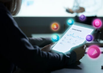 A person holds a tablet displaying a visitor activity graph, while colorful digital icons representing analytics, search, communication, and data surround the scene. Another person is blurred in the background. | FintechZoom