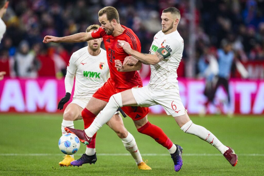 Kane controls the ball while being closely challenged by  two FC Augsburg opponents in white jerseys during FC Augsburg vs. Bayern Munich match, with fans visible in the stadium background. | FintechZoom 