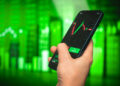 A hand holding a smartphone displaying a candlestick stock chart with buy and sell options. In the background, green financial graphs and charts are visible, suggesting positive market movement. | FintechZoom