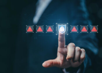 A person in a suit touches a glowing digital shield icon with a lock, surrounded by red warning triangles with exclamation marks, symbolizing security challenges fintech startups face in cybersecurity and data protection. | FintechZoom