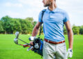 A man in a light blue polo shirt and white pants stands on a golf course, carrying his golf bag over his shoulder, ready for a PGA Tour day. The background shows green grass and trees under a blue sky. | FintechZoom