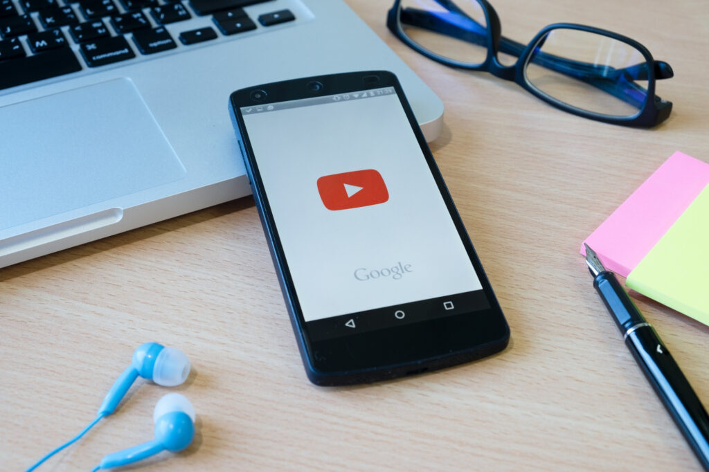 A smartphone displaying the YouTube app sits on a wooden desk near a laptop keyboard, glasses, blue earphones, a pen, and colorful sticky notes—capturing the perfect setup for streaming your favorite YouTube content. | FintechZoom
