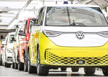 A row of colorful Volkswagen electric vans, led by a yellow and white one, are lined up in a bright, modern factory—a vibrant showcase of innovation driving the Global Electric Vehicle Market forward. | FintechZoom