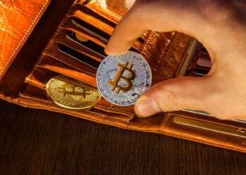 A hand holds a silver Bitcoin coin above an open leather wallet containing cards and a gold Bitcoin coin, all placed on a wooden surface. | FintechZoom