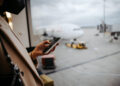 A person in a tan coat holds a smartphone while standing near a large airport window, looking out at a parked airplane on the runway. The scene appears to be indoors, possibly in a departure lounge. | FintechZoom