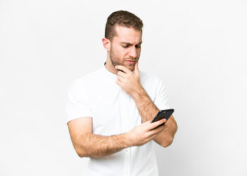 A man with short hair and a beard, wearing a white shirt, stands against a white background, looking at his smartphone with a puzzled expression as he wonders if his Android phone could have stalkerware. | FintechZoom