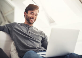 A man with dark hair and a beard sits on a couch, smiling while using his laptop to check if the casino offers a no deposit bonus. He is wearing a gray long-sleeve shirt and appears relaxed in a bright, modern room. | FintechZoom