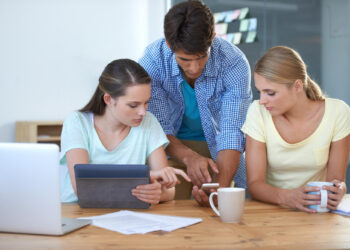 Three young adults collaborate at a desk with a tablet, smartphone, and coffee mugs. Two women sit while a man stands between them, all focused on the devices. Papers and a laptop are on the wooden table. | FintechZoom