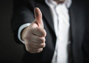 A person wearing a black suit and white shirt gives a thumbs-up gesture, with the hand in focus and the face out of view. | FintechZoom