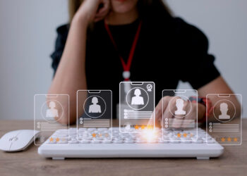 A person sits at a desk typing on a keyboard with holographic icons of user profiles and star ratings above the keys, suggesting digital recruitment or streamlining HR and payroll processes. | FintechZoom