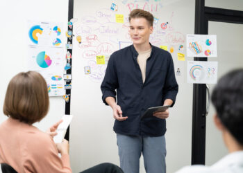 A person stands holding a tablet and presenting an employee training strategy to seated colleagues in an office. Behind them is a whiteboard with colorful charts, diagrams, and sticky notes on business planning and fintech team performance. | FintechZoom