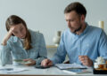 A woman looks stressed while holding cash, as a man calculates with a calculator. Both sit at a table with papers, suggesting they are dealing with debt or financial concerns and budgeting. | FintechZoom