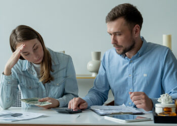 A woman looks stressed while holding cash, as a man calculates with a calculator. Both sit at a table with papers, suggesting they are dealing with debt or financial concerns and budgeting. | FintechZoom