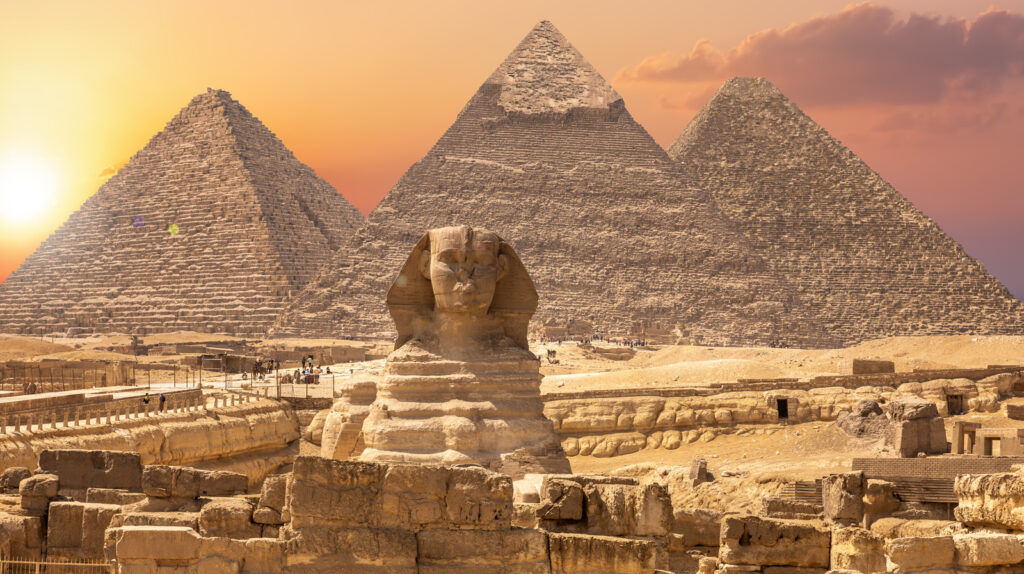 The Great Sphinx of Giza with three pyramids in the background, photographed at sunset with a golden sky and the sun near the horizon. | FintechZoom