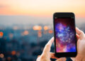 A person holds a smartphone displaying a colorful zodiac wheel with astrological symbols, against a blurred cityscape at sunset. | FintechZoom