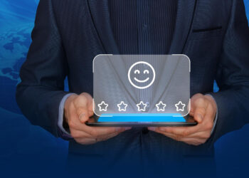 A person in a suit holds a tablet displaying a digital graphic of a smiling face above five stars, symbolizing high customer satisfaction or positive feedback. A faint image of a globe appears in the background. | FintechZoom