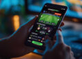 A person holds a smartphone displaying a sports betting app, showing live soccer match updates and betting options, with the “Place Bet” button visible. A computer screen is blurred in the background. | FintechZoom