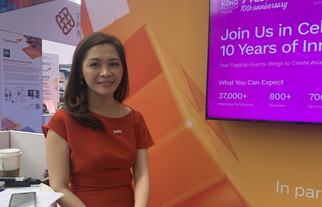 Daisy Ip in a red dress smiles while sitting at a booth during Hong Kong FinTech Week. Behind her is a purple screen displaying event details and statistics about attendees, speakers, and sponsors. | FintechZoom