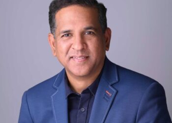 Shiva Ramnarine, a man with short dark hair, wearing a navy blue suit jacket over a dark shirt, smiles at the camera against a plain light gray background. | FintechZoom