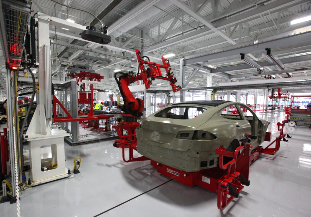 A robotic arm assembles a car body on an automated production line inside a bright, modern automotive factory with metal beams and high ceilings. | FintechZoom