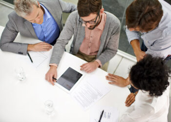 Four people sit and stand around a white table, looking at documents and a tablet while discussing Choosing the Best AML Software. Pens, papers, and glasses of water are also on the table during this business meeting. | FintechZoom