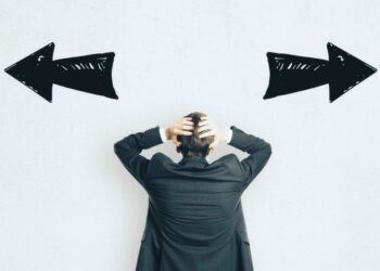 A person in a black suit holds their head in frustration, standing between two large black arrows pointing left and right on a white background, symbolizing indecision or choice. | FintechZoom