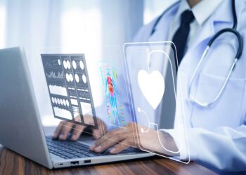 A doctor in a white coat uses a laptop, with digital medical icons and data holograms, including a heart symbol, representing modern healthcare technology advances for PTC Therapeutics' rare-disease solutions. | FintechZoom