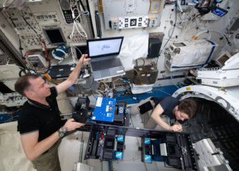 Two astronauts work inside a space station module, surrounded by equipment, wires, and control panels. One holds a laptop, while the other adjusts a device on a large blue apparatus. | FintechZoom
