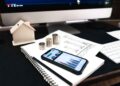 A smartphone displaying colorful financial graphs rests on a spiral notebook beside coins, a house model, and documents—everything you need to Build Your Own Finance Tracker—on a desk with a computer monitor in the background. | FintechZoom