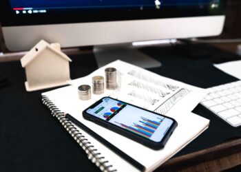 A smartphone displaying colorful financial graphs rests on a spiral notebook beside coins, a house model, and documents—everything you need to Build Your Own Finance Tracker—on a desk with a computer monitor in the background. | FintechZoom