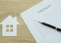 A paper checklist and a pen lie on a wooden surface next to a white paper cutout of a house, suggesting tasks or items related to home ownership or real estate. | FintechZoom