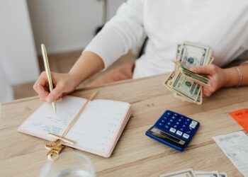 Person sitting at a desk holding cash, writing in a planner with a pen. A blue calculator, receipts, and a glass of water are on the wooden table. Only the person's torso and hands are visible. | FintechZoom