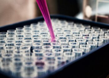 A close-up of a pink liquid being dispensed from a pipette into a tray filled with multiple clear plastic test tubes, suggesting a laboratory or scientific experiment setting. | FintechZoom