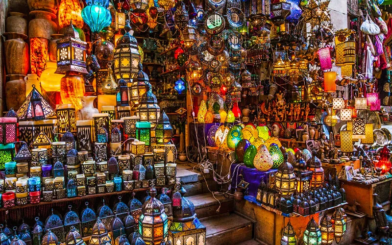 A vibrant market stall displays colorful, intricately designed lanterns and lamps, glowing in a variety of patterns and shapes, stacked on shelves and hanging from above, creating a warm and lively atmosphere. | FintechZoom