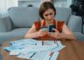 A woman sits on a couch, looking concerned while holding credit cards and a phone. Many bills and financial documents are spread out on the table in front of her. | FintechZoom
