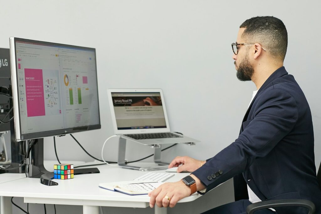 A man wearing glasses and a blazer works at a desk with two monitors, a notebook, and a Rubik's cube. He is focused on his screens, one displaying fintech charts and slides, the other a website. | FintechZoom