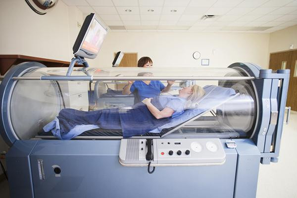 A patient lies inside a Hyperbaric Chamber in a medical facility, covered with a blue blanket, while a healthcare worker stands nearby talking and monitoring. | FintechZoom