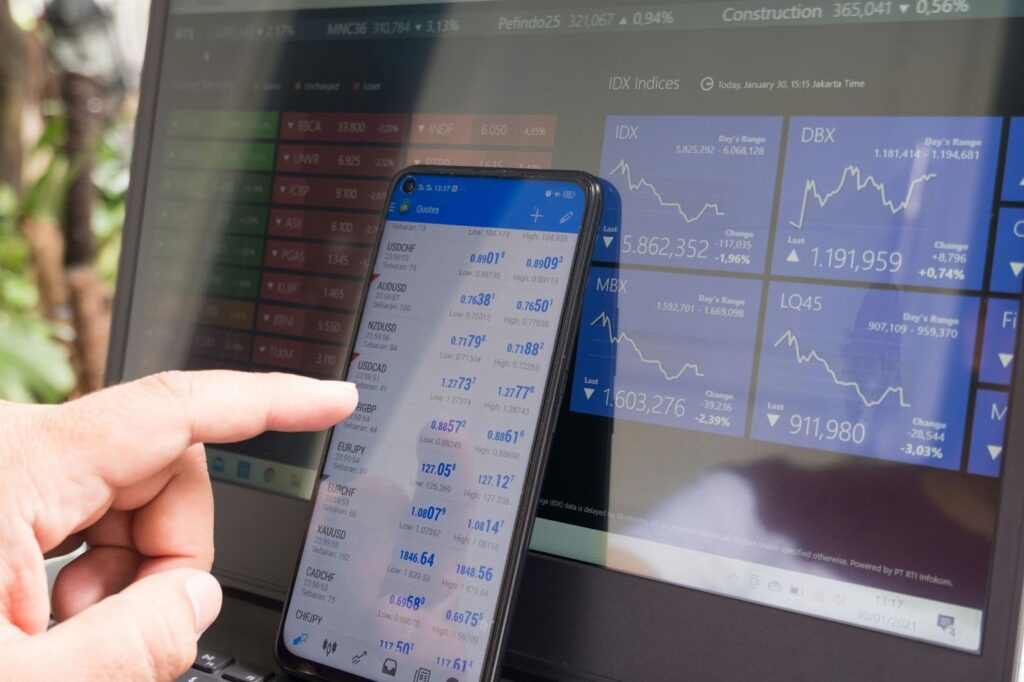 A hand points at a smartphone displaying fintech-powered currency exchange rates, while a laptop in the background shows financial data and stock market indices. | FintechZoom