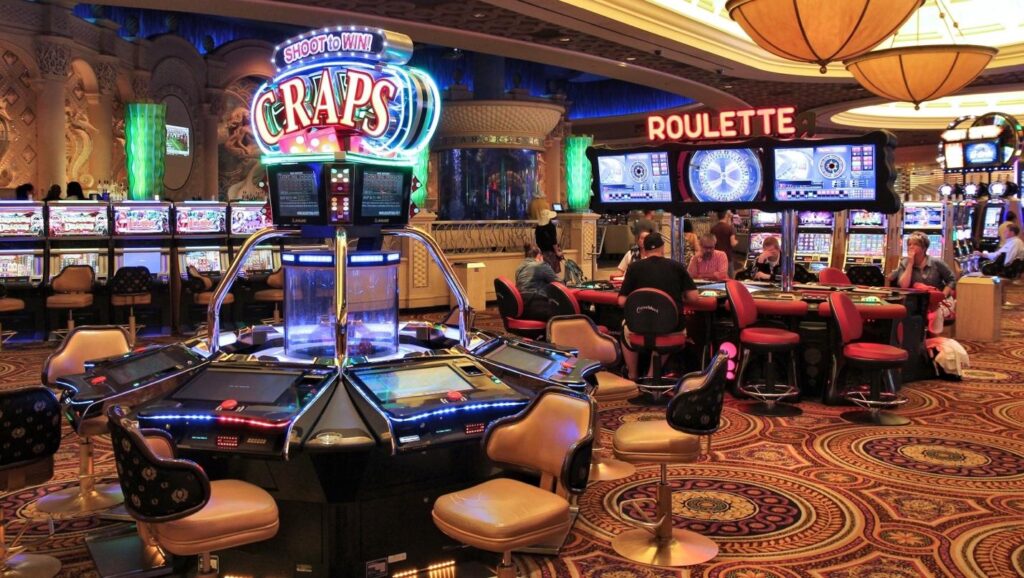A lively casino interior with electronic craps and roulette tables, bright neon signs, slot machines in the background, and people sitting and playing on various gaming tables under ornate ceiling lights. | FintechZoom