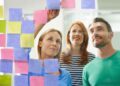 Three people stand in front of a glass wall covered with colorful sticky notes, collaborating and smiling as one person places a note on the wall—perfect for brainstorming and onboarding teams. | FintechZoom