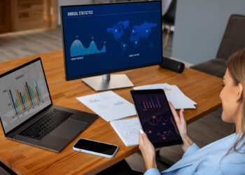 A woman sits at a desk analyzing charts and graphs from Modern Asset Management Platforms displayed on a laptop, tablet, and monitor. Papers with printed graphs, a smartphone, and a pen highlight the data-focused work environment. | FintechZoom