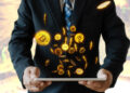 A person in a suit holds a tablet, with glowing Bitcoin symbols and coins floating above, symbolizing digital currency and cryptocurrency transactions. Cryptocurrencies are becoming popular in the world of betting. | FintechZoom