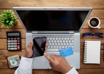 A person holds a smartphone and a credit card above a laptop on a wooden desk with a calculator, cash, a plant, coffee, sunglasses, a notebook, and pens. | FintechZoom