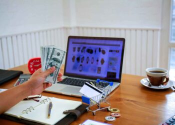 A hand holding several US dollar bills is in front of a laptop displaying an online shopping website. A notebook, a cup of coffee, a clock, a small shopping cart with cards, and some coins are on the wooden desk. | FintechZoom