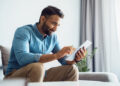 A man with a beard, wearing a blue shirt and khaki pants, sits on a sofa and smiles while using a tablet in a bright, modern living room. | FintechZoom