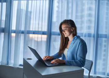 A woman wearing glasses and a blue shirt sits at a desk, smiling while managing her bank account on a laptop. Large windows with sheer curtains and city buildings are visible in the background. | FintechZoom