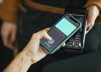 A person holds a smartphone near a payment terminal, using a mobile wallet app and Digital Payment Solutions to make a contactless payment. Another person holds the terminal steady during the transaction. | FintechZoom