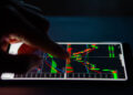 A person uses their finger to interact with a smartphone displaying colorful financial candlestick charts, indicating active stock trading or cryptocurrency market data on a dark background. | FintechZoom
